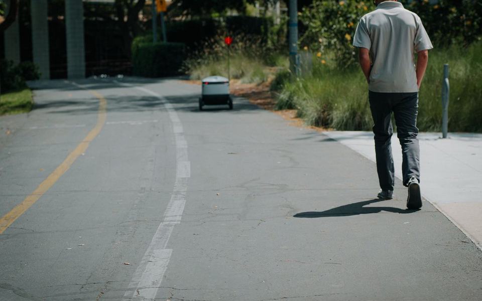 Man seen from behind walking along a road edge while a small delivery robot appr