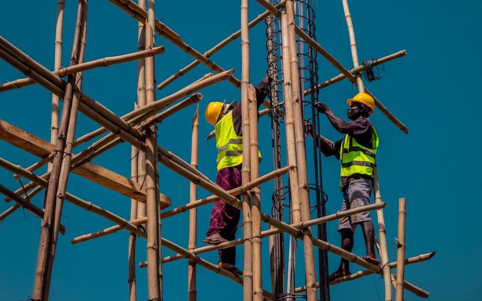 Two workers in hi-vis vests and hard hats climbing high on bamboo scaffolding ag