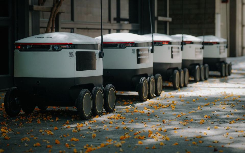 A line of five or six identical delivery robots parked/travelling in a row besid