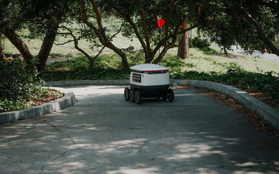Autonomous delivery robot travelling along a curved paved path under trees, clea