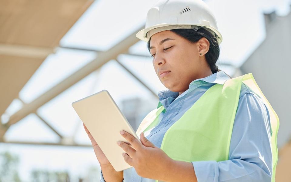 Woman in white hard hat and hi-vis vest looks down at a tablet on a site under s