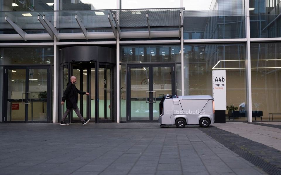Pedestrian walks past a revolving door while a small autonomous cleaning robot s