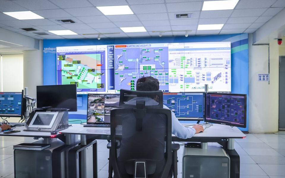 Operator seen from behind at a desk of monitors facing a video wall in a bright